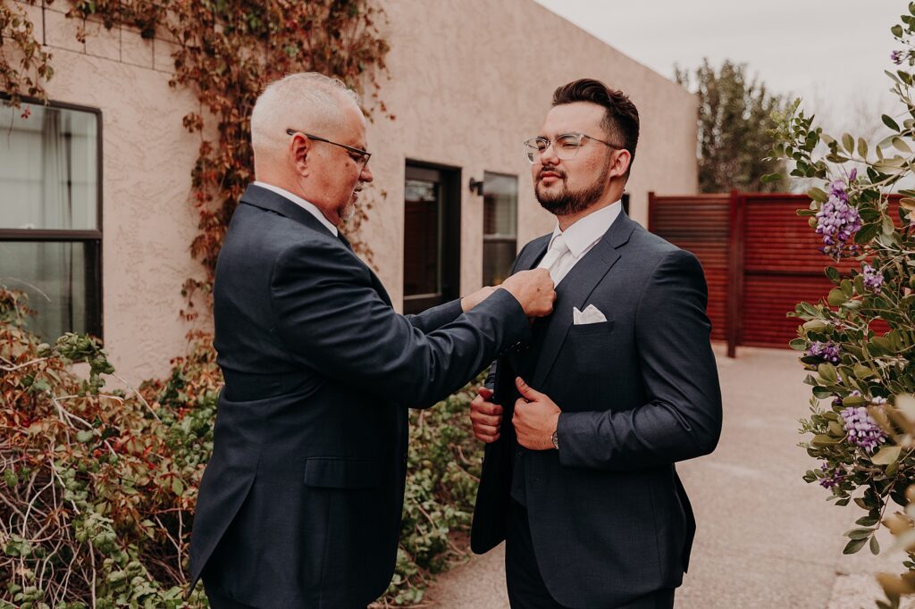 father helps groom put on suit and jacket on wedding day