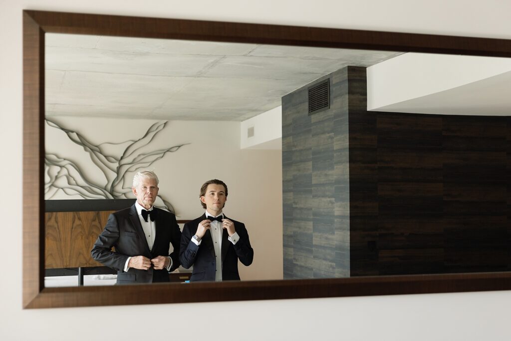 groom and his father adjusting bowties in mirror in hotel suite