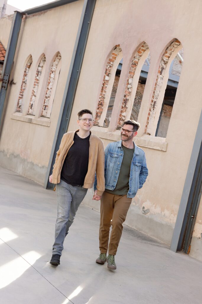 LGBTQAI+ couple walk together smiling in front of stone arches at Monroe Street Abbey in Phoenix.