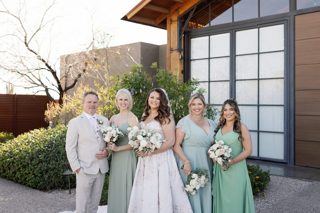 Bride and wedding party in green dresses smile while holding bouquets.