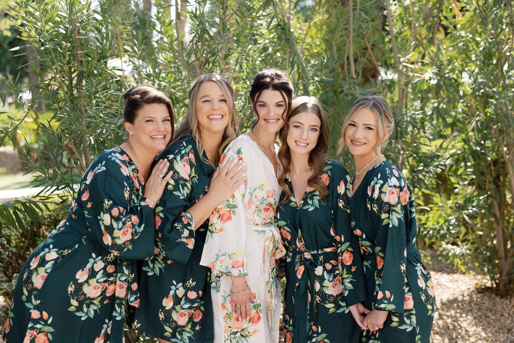 Bride and bridesmaids hug and smile wearing matching robes in front of palm trees at the Scott.