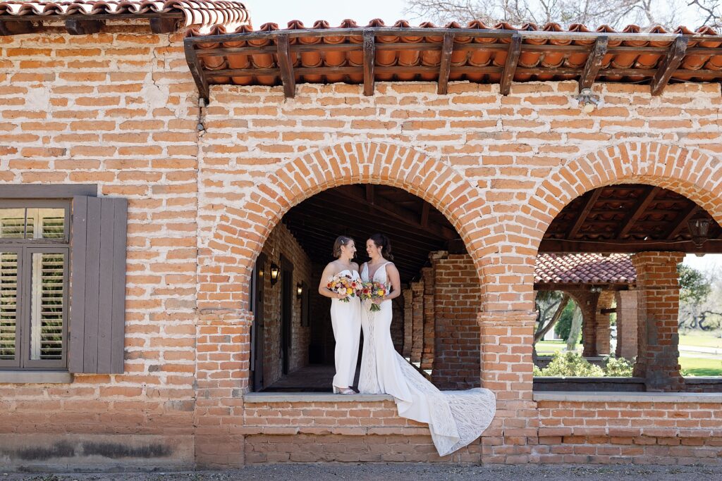 Two brides stand together under red brick archway on wedding day at the Tubac Resort and Spa.