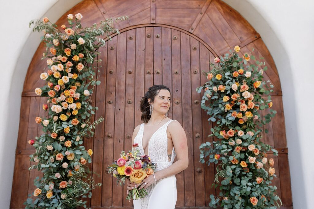 Bride looks over shoulder holding colorful bouquet in front of wooden doors and floral arch.