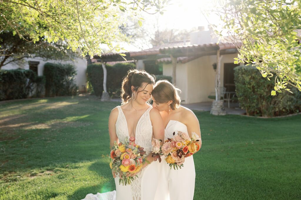 Bride kisses her wife's shoulder in warm sunlight in grassy courtyard.