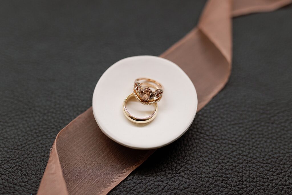 wedding rings lay in white ceramic dish with mauve ribbon styled underneath